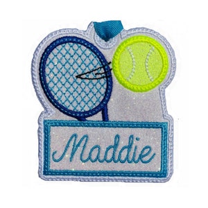 Könnte beinhalten: Ein weißer Glitzer-Tennis-Anhänger mit einem blauen Tennisschläger, einem gelben Tennisball und dem Namen "Maddie" in blauer Kursivschrift.