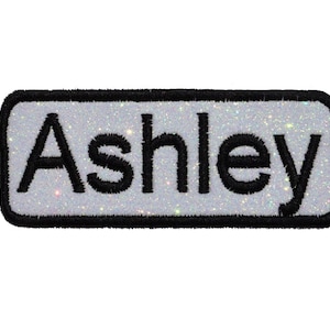 May include: A white, sparkly, rectangular iron-on patch with a black border and the name "Ashley" in black lettering.