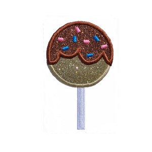 Op de afbeelding: Een bruine en gouden glitter cake pop met blauwe en roze sprinkles erop. De cake pop zit op een witte stok.