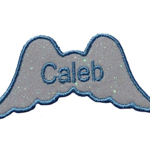 Puede incluir: Un parche de ala de ángel de brillantina blanca con un contorno azul y el nombre "Caleb" bordado en hilo azul.
