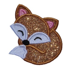 May include: A fox-shaped embroidered patch featuring a brown glittery finish, light blue accents on the face and ears, pink inner ear details, and a small black nose. The design shows a stylized fox head with closed eyes and a stitched border.