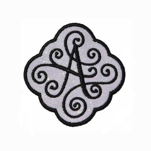 Peut inclure: Un écusson thermocollant blanc pailleté avec un contour noir en forme de diamant. L'écusson présente une lettre "A" noire stylisée avec des volutes autour.