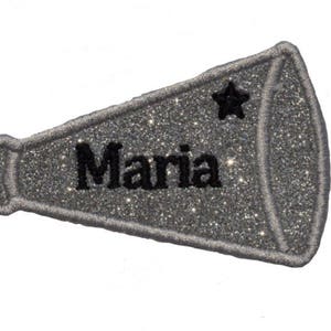 May include: A silver glitter megaphone patch with the name "Maria" in black block letters. A black star is above the name. The patch has a gray stitched border and is on a white background.