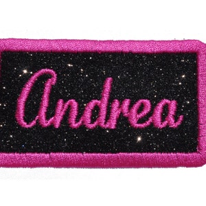 May include: Pink glitter iron-on patch with the name "Andrea" embroidered in pink on a black background.
