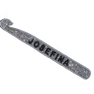 May include: A silver glitter crochet hook with the name "JOSEFINA" in black lettering. The hook is a crafting tool, likely used for crocheting, and is set against a white background.