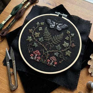 Puede incluir: Un bastidor de bordado redondo de madera con tela negra que presenta un diseño bordado a mano de una polilla, enredaderas de hiedra verde, helechos y setas rojas con puntos blancos, colocado sobre tela negra junto a hilo de bordar y tijeras.