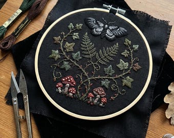 May include: A round wooden embroidery hoop containing black fabric with a hand-stitched design of a moth, green ivy vines, ferns, and red mushrooms with white spots. The hoop is placed on black fabric next to two skeins of embroidery floss and metal scissors.