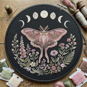 May include: Embroidered moth and floral design on a black fabric hoop. The moth is in shades of pink and brown, surrounded by flowers and phases of the moon. Embroidery threads in various colours are visible around the hoop.