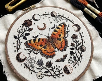 May include: An embroidery hoop containing a fabric piece with a central orange and black butterfly surrounded by embroidered motifs including moon phases, spider webs, small skulls, bats, and thorny rose stems in black and orange thread.