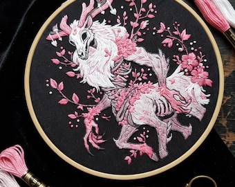 May include: A circular wooden embroidery hoop containing black fabric with a detailed thread-stitched design of a skeletal deer creature with pink antlers, fur, and floral accents. Includes two needles, gold-toned scissors, and pink and white thread.