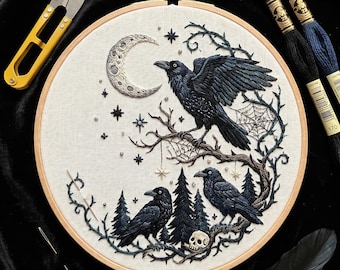 May include: An embroidery hoop featuring a detailed needlework design of three black ravens perched on thorny branches among pine trees and a skull. A crescent moon and stars are stitched above, with a spiderweb accent. Yellow scissors and embroidery floss are nearby.