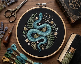 May include: A circular wooden embroidery hoop containing black fabric with a teal snake stitched in the center, surrounded by embroidered green ferns and small flowers. The hoop is arranged with embroidery scissors, thread, and books.