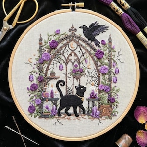 May include: An embroidery hoop containing a fabric panel with a detailed needlework design of a black cat standing before a gothic archway. The scene features purple roses, a black raven, spiderwebs, candles, crystals, and a small cauldron.