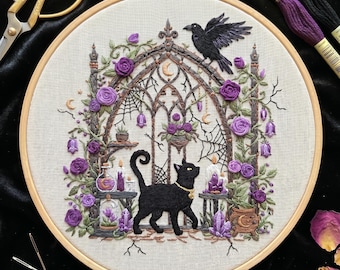 May include: A round wooden embroidery hoop containing a fabric panel with a detailed needlework design of a black cat, a raven, purple roses, and a gothic archway. The scene includes crystals, candles, and spiderwebs, with gold scissors and thread nearby.