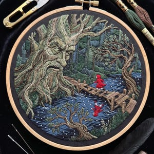May include: A circular wooden embroidery hoop containing a detailed thread-stitched scene of a large, ancient tree with a face overlooking a small figure in a red cloak crossing a wooden bridge over a dark blue river, with embroidery tools nearby.