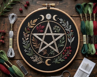 May include: A circular wooden embroidery hoop containing black fabric with a stitched pentagram surrounded by a wreath of leaves, berries, and moon phases. The kit includes multiple skeins of embroidery floss in green, red, and silver, two pairs of scissors, and a pack of needles.