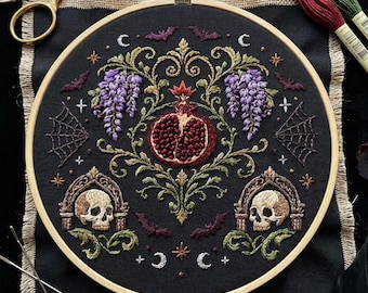 May include: A circular embroidery hoop containing black fabric with a hand-stitched design featuring a central pomegranate, two skulls in arched frames, purple wisteria, spiderwebs, bats, and crescent moons, accompanied by gold scissors.