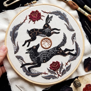 Op de afbeelding: Een ronde houten borduurring met crèmekleurige stof en handgeborduurde ontwerpen. Het borduurwerk toont een zwart konijn en een zwarte wolf, beide met spinnenwebpatronen, omringd door twee donkerrode rozen en grijze veren.