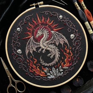Könnte beinhalten: Ein runder Stickrahmen mit einem schwarzen Stoffeinsatz, der ein handgesticktes Motiv eines grauen Drachen vor einer roten Sonne zeigt. Der Drache ist von orangefarbenen und roten Flammen, dornigen Ranken und kleinen weißen Totenköpfen umgeben.