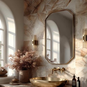 May include: A gold-toned oval vessel sink sits on a brown marble countertop. Above the sink is a wall-mounted gold faucet and a rectangular mirror with rounded corners. Two gold wall sconces flank the mirror, and a vase with dried flowers sits nearby.