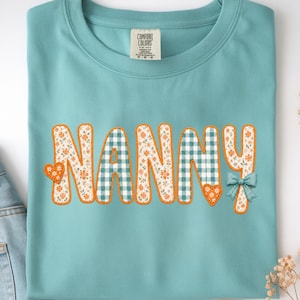 May include: A teal crewneck t-shirt featuring the word NANNY in large letters. The letters alternate between a white floral pattern with orange flowers and a teal and white gingham check pattern. Two orange hearts and a small teal bow are included.