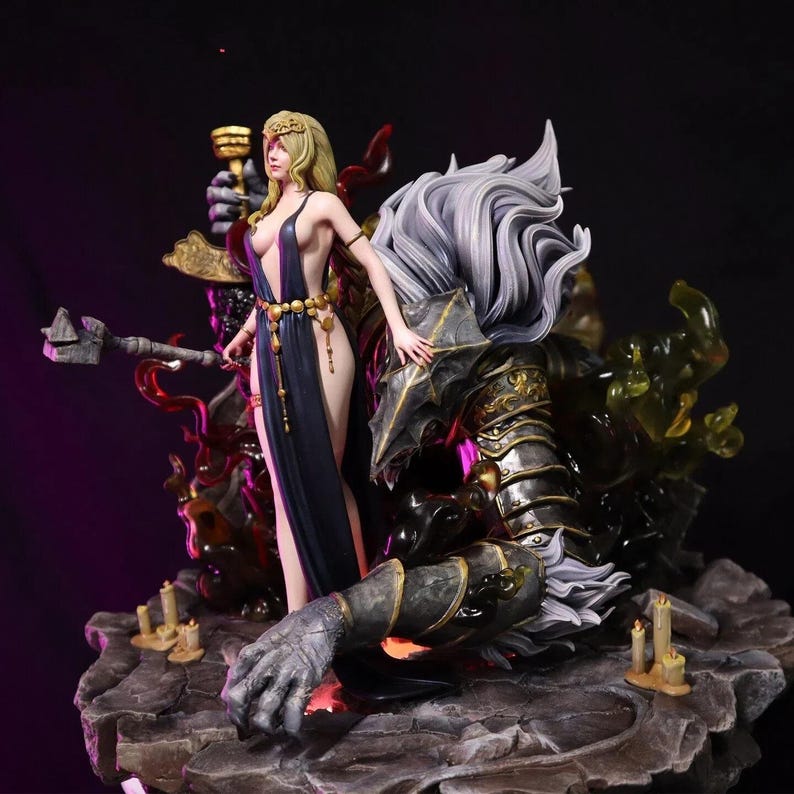 May include: A detailed figurine depicting a woman in a flowing dark blue dress, holding a chalice, standing near a fallen armored figure. The statue features intricate details, with a stone base and candle accents. The woman has blonde hair and a gold headpiece.