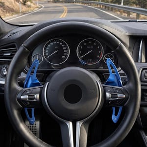 Puede incluir: Un volante de cuero negro con levas de cambio de aluminio azul brillante con símbolos de más y menos en blanco. El volante está situado frente a un salpicadero con dos indicadores analógicos circulares y una vista de la carretera.