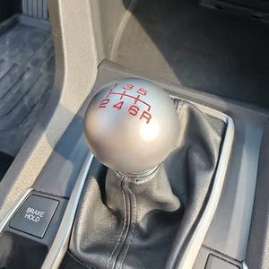 May include: A spherical silver gear shift knob with a six-speed manual transmission pattern engraved in red, featuring numbers one through six and an R for reverse, mounted on a black leather boot within a car centre console with a brake hold button.