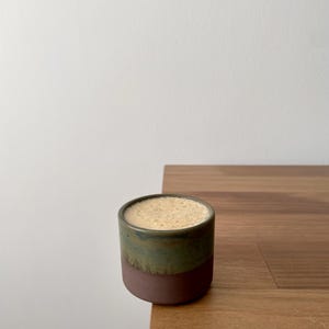 May include: A cylindrical ceramic cup with a two-tone glaze, featuring a dark brown matte base and a mottled olive green upper section. The cup is filled to the brim with a light tan, frothy beverage with a fine, bubbly texture on the surface.