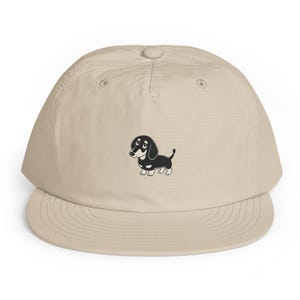 Puede incluir: Una gorra de béisbol de color beige con una ilustración gráfica centrada de un perro salchicha en blanco y negro. La gorra presenta una corona estructurada, una visera plana y ojales cosidos en los paneles superiores del diseño.
