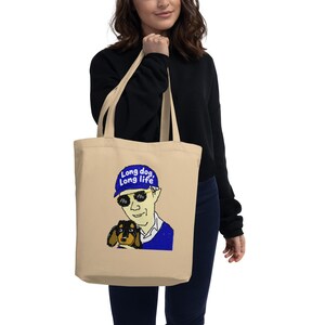 Puede incluir: Una bolsa de tela beige con una ilustración gráfica de una persona que lleva una gorra azul con el texto Long dog, Long life, gafas de sol oscuras y un suéter azul, sosteniendo a un pequeño perro salchicha de color negro y fuego.