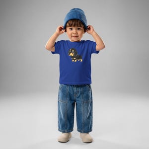 Könnte beinhalten: Ein blaues Kurzarm-T-Shirt mit einer Grafik eines schwarz-lohfarbenen Dackels, kombiniert mit einer blauen Strickmütze und einer blauen Jeans. Das T-Shirt zeigt eine cartoonartige Illustration des Hundes auf der vorderen Brustpartie.