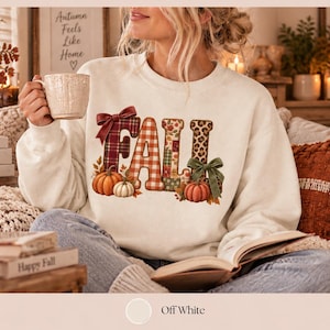 Könnte beinhalten: Ein cremefarbenes Sweatshirt mit Rundhalsausschnitt und dem Wort FALL in großen, gemusterten Buchstaben. Das F ist rot kariert mit einer Schleife, das A orange kariert, das L floral und das L mit Leopardenmuster und einer grünen Schleife. Kürbisse liegen am Fuß.