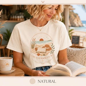 Könnte beinhalten: Cremefarbenes T-Shirt mit einem kreisförmigen Aufdruck, der eine Strandszene und den Text "Beach Book Club" zeigt. Das Wort "Natural" steht unten. Die Person trägt das T-Shirt und hält ein Buch, mit einer Kaffeetasse und Strand im Hintergrund.