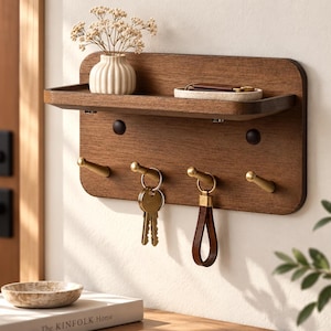 May include: A rectangular wooden wall-mounted organizer featuring a top shelf, four brass-toned hooks, and two dark circular accents. The shelf holds a small white ribbed vase with dried flowers and a white tray. Keys hang from the brass hooks.