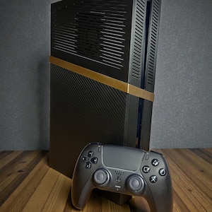 Op de afbeelding: Een zwarte spelcomputer met een horizontale gouden band in het midden, een onderkant met een koolstofvezeltextuur en een ventilatierooster aan de bovenkant. Een bijpassende zwarte draadloze controller met twee analoge sticks staat ervoor.