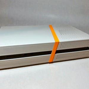 May include: A rectangular white gaming console shell featuring a two-piece design held together by a bright orange elastic band. The top surface has a series of parallel ventilation slits, and the side panels also feature thin, vertical vent lines.