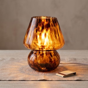 Puede incluir: Una lámpara de mesa con forma de seta hecha de cristal de color ámbar con un patrón moteado marrón oscuro. La lámpara presenta una pantalla ancha y acampanada y una base redondeada, emitiendo una luz cálida.