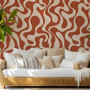 May include: A low-profile woven rattan sofa featuring a white seat cushion and an assortment of cream, white, and striped throw pillows. The sofa is positioned against a wall covered in wallpaper with a bold, wavy, terracotta and cream pattern.