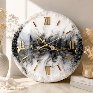 May include: A round wall clock featuring a white and black marbled resin face with gold-colored Roman numerals and ornate hands. The outer edge is decorated with a textured border of clear and black crushed glass-like stones.