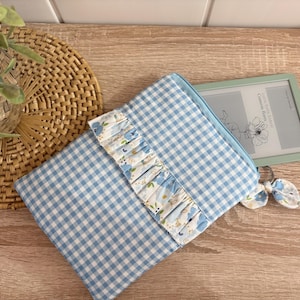 May include: A light blue and white gingham fabric sleeve for an e-reader, featuring a horizontal ruffled band with a blue floral pattern. A small matching fabric bow charm is attached to the zipper pull, and a digital e-reader is partially visible.