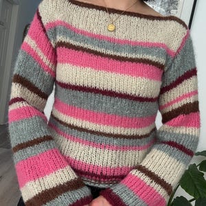 Könnte beinhalten: Ein gestreifter Pullover mit U-Boot-Ausschnitt und langen Ärmeln. Der Pullover hat horizontale Streifen in Rosa, Grau, Braun und Creme. Die Ärmel sind ausgestellt. Der Pullover ist aus weichem Strickmaterial gefertigt.