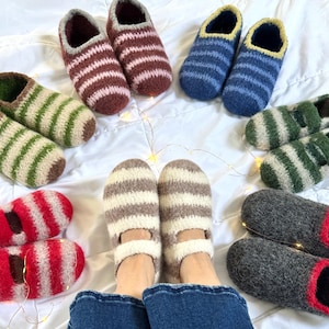 Puede incluir: Una variedad de zapatillas de lana a rayas hechas a mano en varios colores, incluyendo rojo, verde, marrón, azul y gris. Las zapatillas están dispuestas sobre una superficie blanca, con un par puesto. Las zapatillas parecen acogedoras y cálidas.