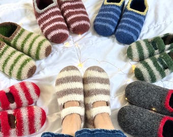 Puede incluir: Una variedad de zapatillas de lana a rayas hechas a mano en varios colores, incluyendo rojo, verde, marrón, azul y gris. Las zapatillas están dispuestas sobre una superficie blanca, con un par puesto. Las zapatillas parecen acogedoras y cálidas.
