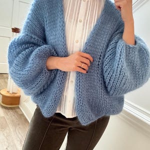 Può includere: Un cardigan azzurro aperti sul davanti con maniche voluminose. Il maglione a maglia ha una consistenza soffice e viene indossato sopra una camicetta bianca. Il cardigan è di colore azzurro pastello.