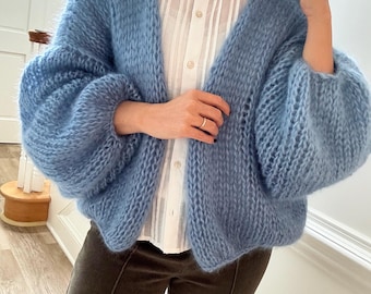 Può includere: Un cardigan azzurro aperti sul davanti con maniche voluminose. Il maglione a maglia ha una consistenza soffice e viene indossato sopra una camicetta bianca. Il cardigan è di colore azzurro pastello.