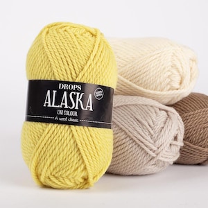 May include: A close-up of several skeins of Drops Alaska Uni Colour yarn. The foreground features a bright yellow skein with a black label. Other skeins are cream, light brown, and tan. The yarn is 100% wool.