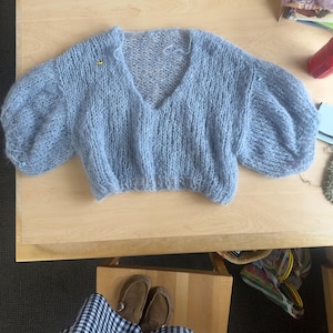 May include: A light blue knitted sweater with a v-neck and puffed sleeves. The sweater is laid flat on a wooden surface.