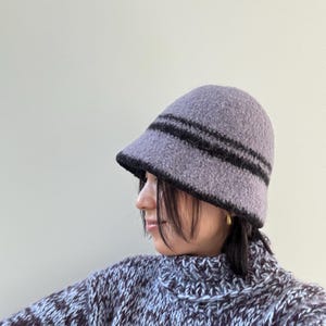 May include: A lavender bucket hat with two black stripes and a black brim. The hat is made of a textured material. The person in the image is also wearing a patterned sweater.