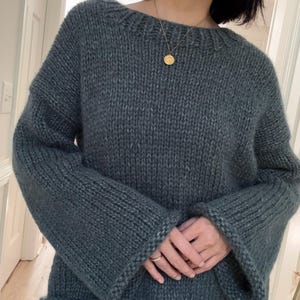 Könnte beinhalten: Ein teal-farbener, grob gestrickter Pullover mit weitem Ausschnitt und ausgestellten Ärmeln. Der Pullover ist aus dickem Garn gefertigt, was ihm ein strukturiertes Aussehen verleiht. Die Ärmel sind lang und enden in einer ausgestellten Manschette. Der Pullover wird mit hellblauen Jeans getragen.